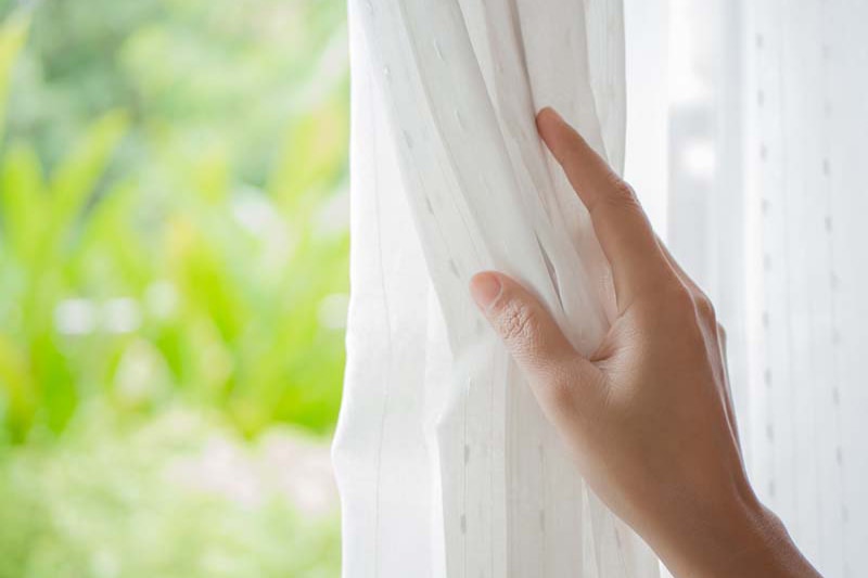 Spring Money-Saving Tips, Woman's hand opening curtains in the bedroom with natural light and garden background.